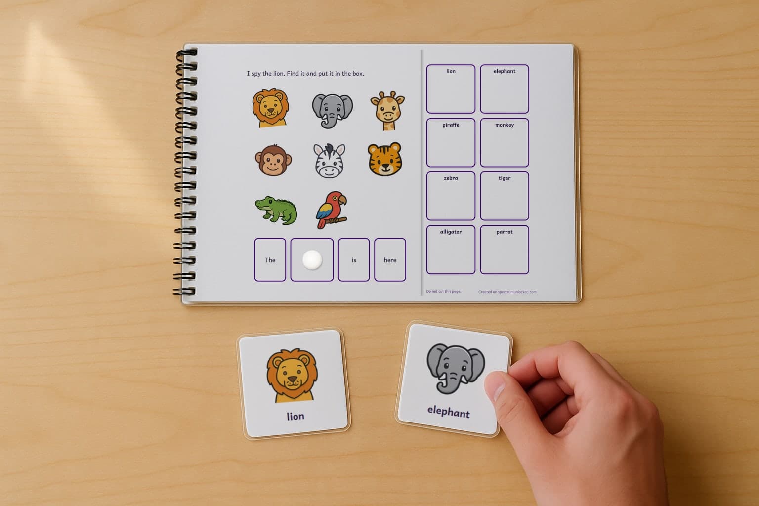 A page of the I Spy Zoo Animals adapted book, laminated and spiral bound, with a child's hand holding a picture card beside it
