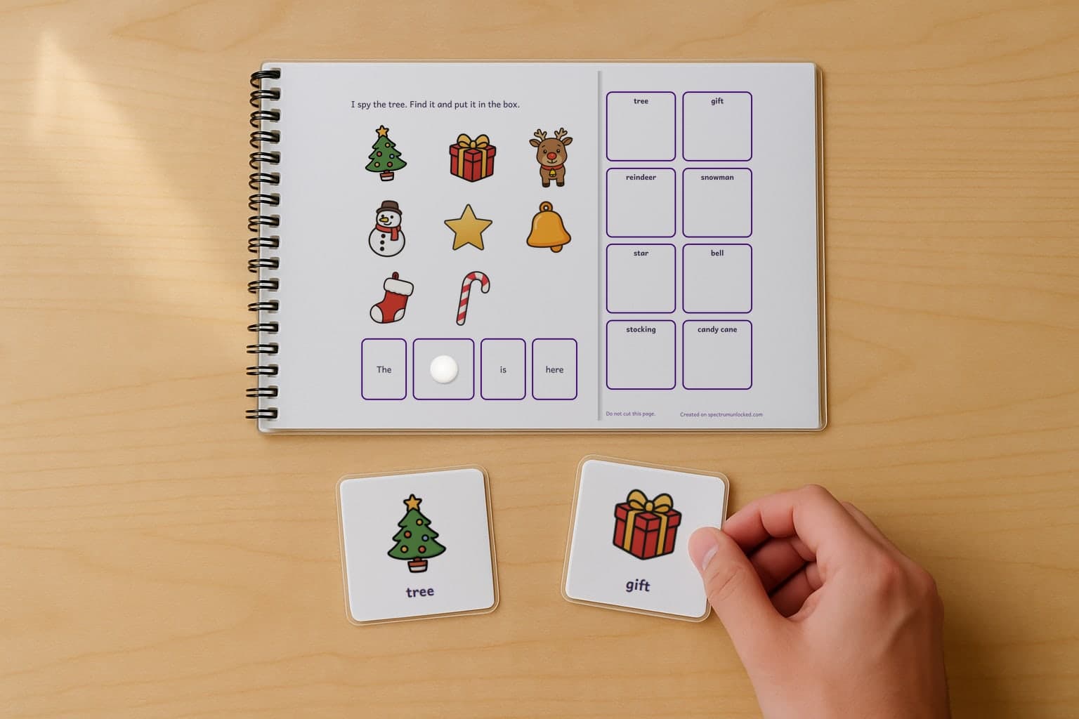 A page of the I Spy Christmas adapted book, laminated and spiral bound, with a child's hand holding a picture card beside it
