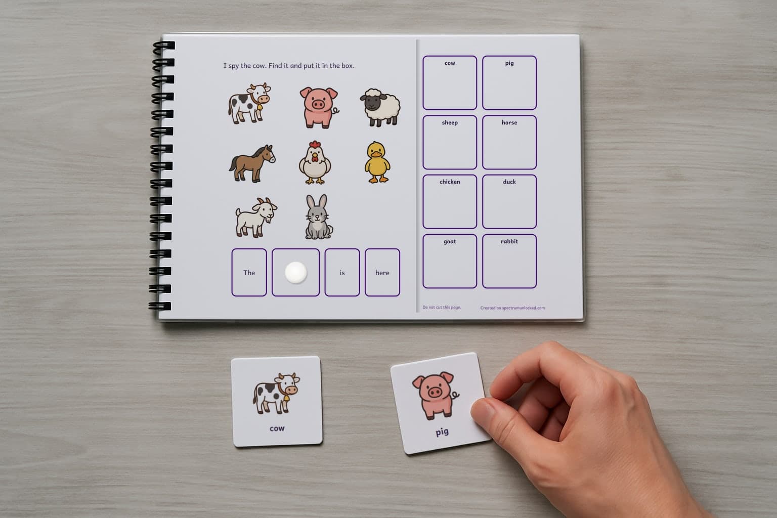 A page of the I Spy Farm Animals adapted book, laminated and spiral bound, with a child's hand holding a picture card beside it