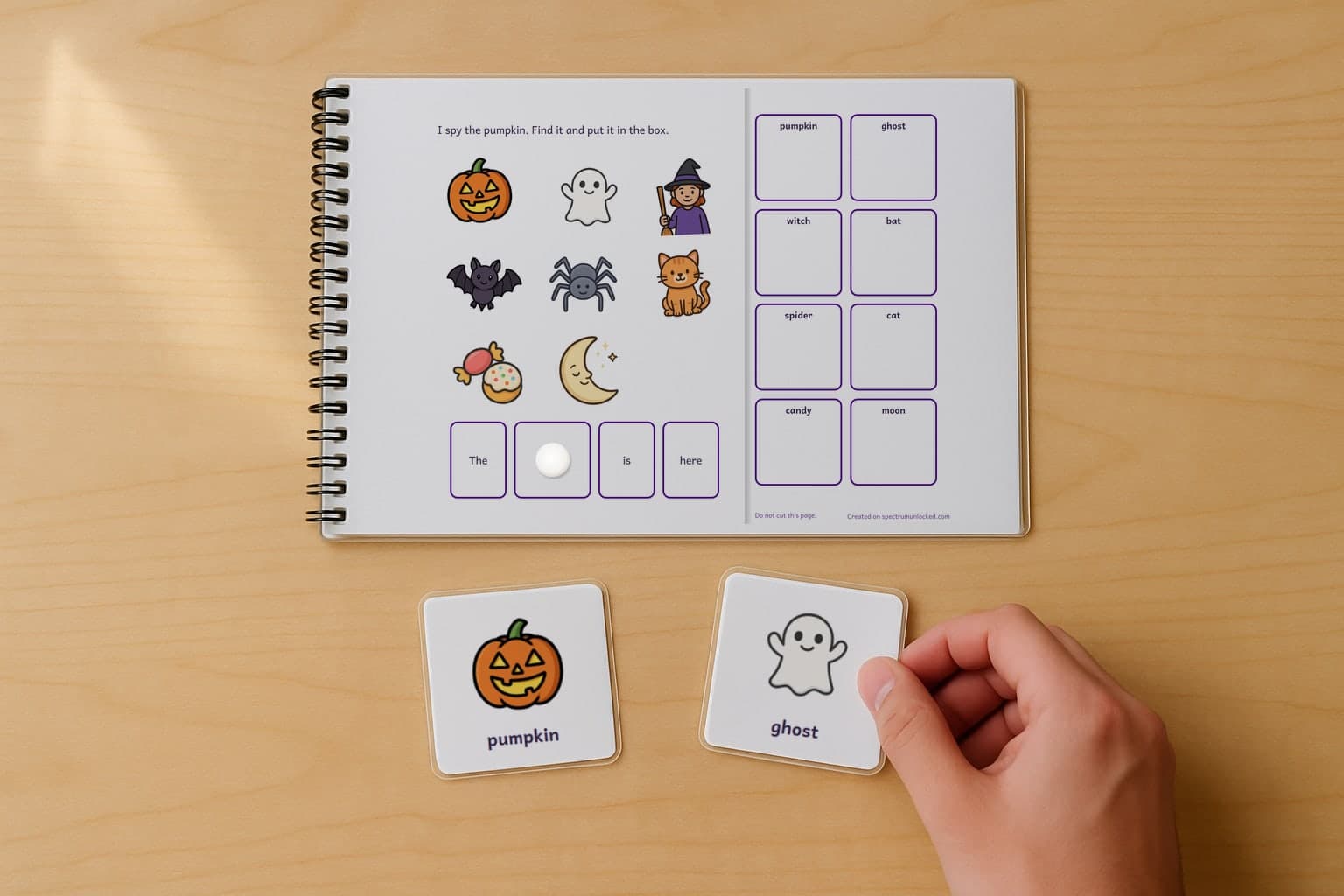 A page of the I Spy Halloween adapted book, laminated and spiral bound, with a child's hand holding a picture card beside it
