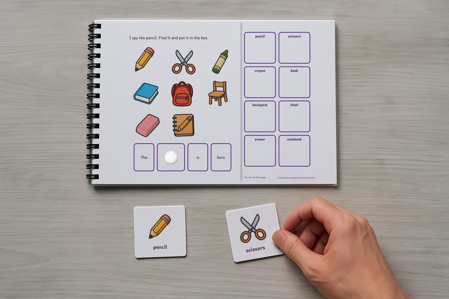 A page of the I Spy Classroom Objects adapted book, laminated and spiral bound, with a child's hand holding a picture card beside it