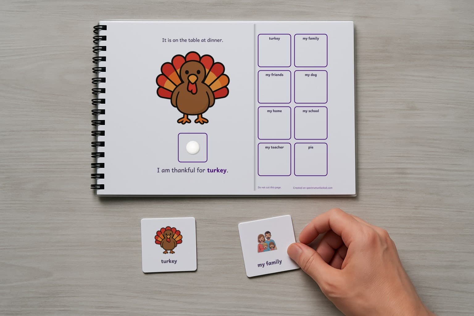 A page of the I Am Thankful adapted book, laminated and spiral bound, with a child's hand holding a picture card beside it