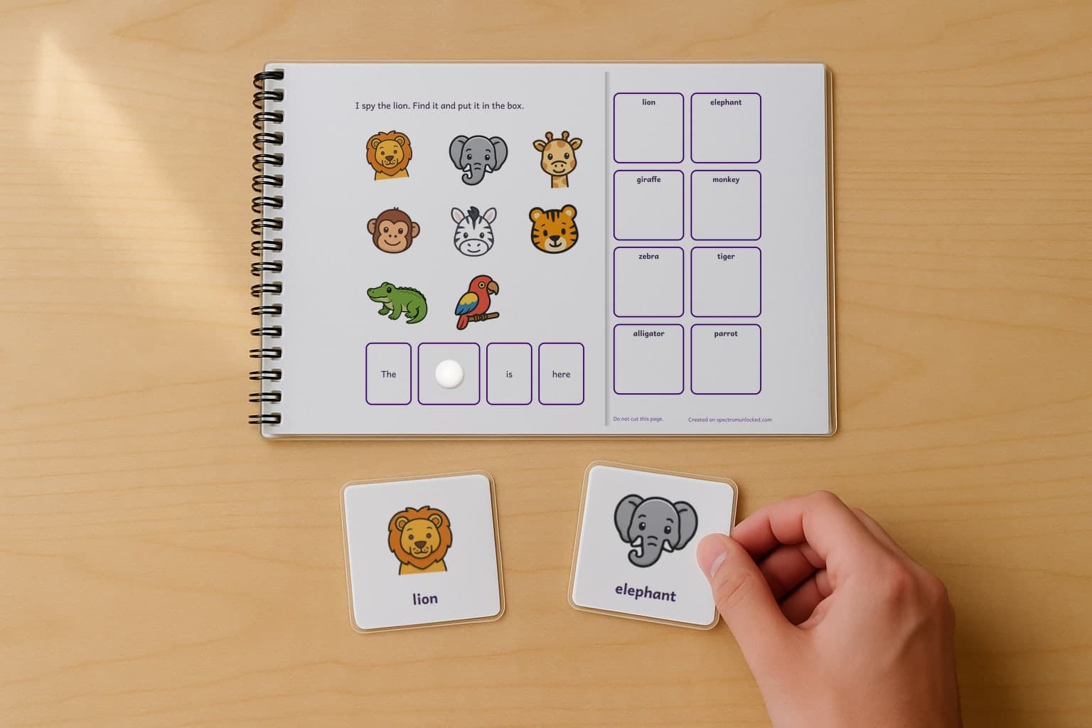 A page of the I Spy Zoo Animals adapted book, laminated and spiral bound, with a child's hand holding a picture card beside it