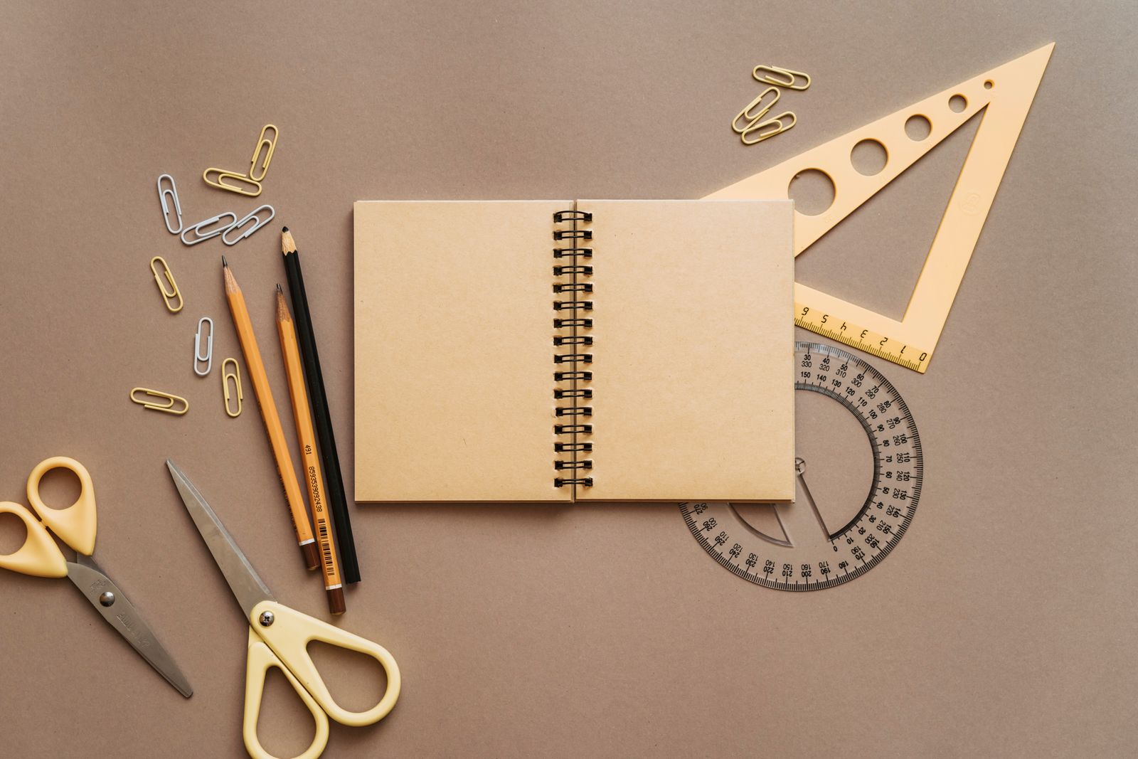 A flat lay of a spiral notebook, pencils, scissors, paper clips, and a triangle ruler on a desk, styled for a back-to-school classroom wishlist
