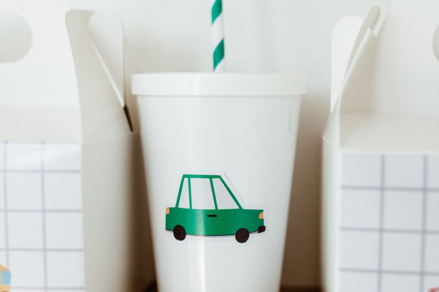 A child's white cup with a green car sticker and a striped paper straw, standing on a wooden shelf between two paper lunch boxes, with no one in the frame