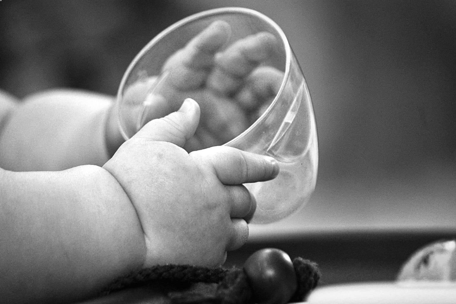 A baby's two hands wrapped around a small clear open cup, photographed in black and white, with no face in the frame