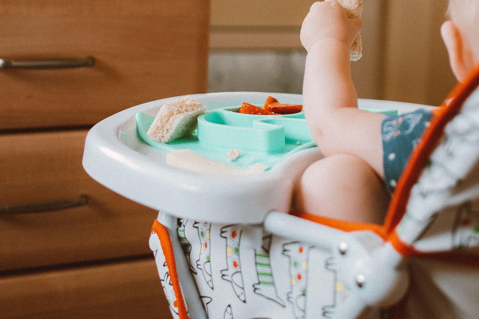 A toddler in a high chair seen from behind in a kitchen, one hand raised with a piece of bread above the tray, the face not visible