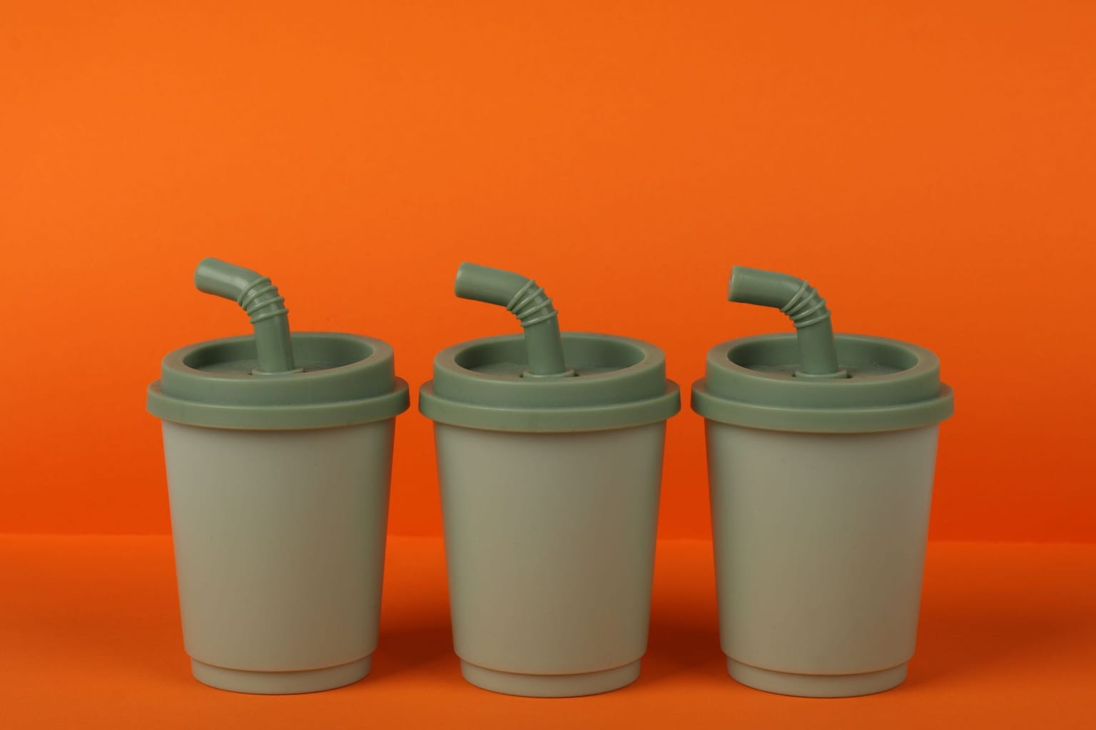 Three green plastic children's cups with bent straws in their lids, standing in a row against an orange background