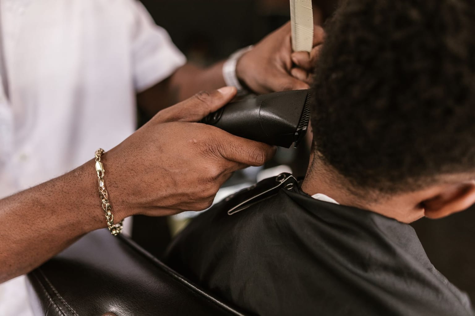 A barber's hands running a small clipper up the back of a child's neck in a barbershop, the child wearing a cape and seen only from behind