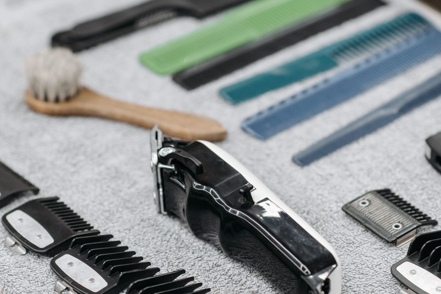 A clipper kit laid out on a gray towel: a cordless clipper, a row of snap-on guide combs, a cleaning brush and several cutting combs, with no person in the frame
