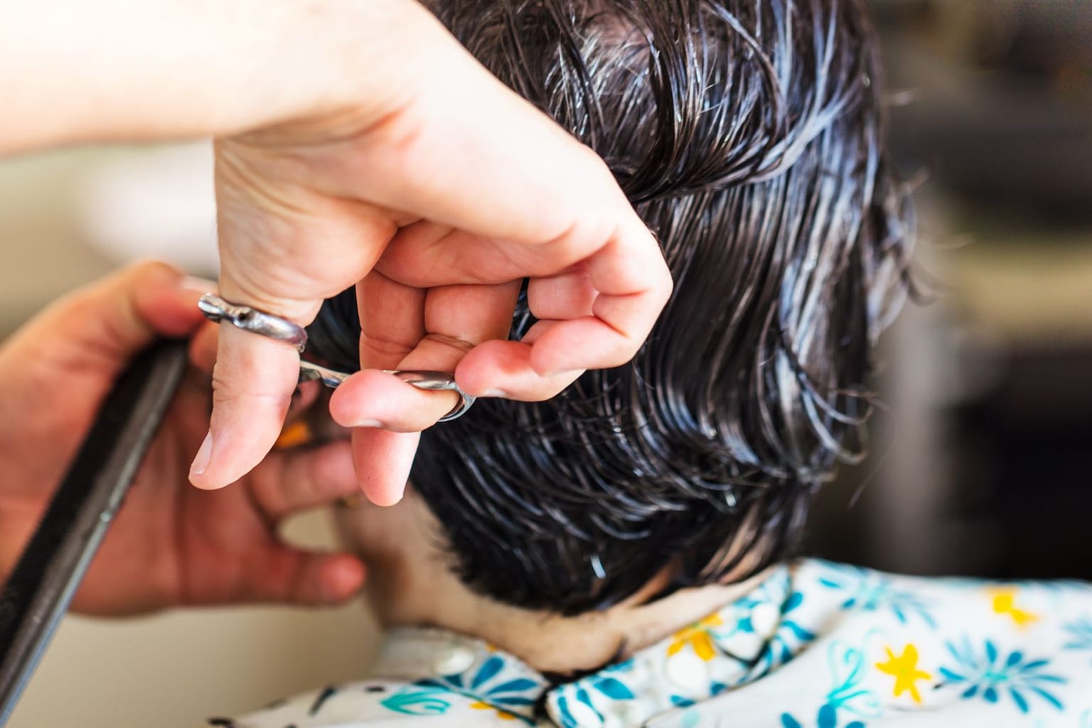 A child seen from behind getting a scissor haircut, an adult's hands holding scissors and a comb against the child's dark wet hair, the child's face not visible