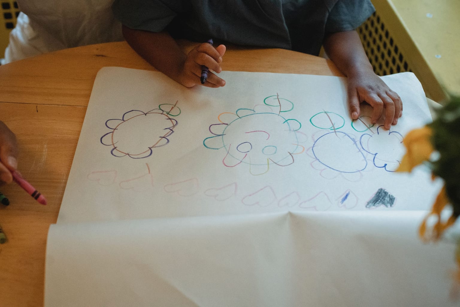 Overhead view of a child drawing crayon flowers on a large sheet of paper at a wooden table