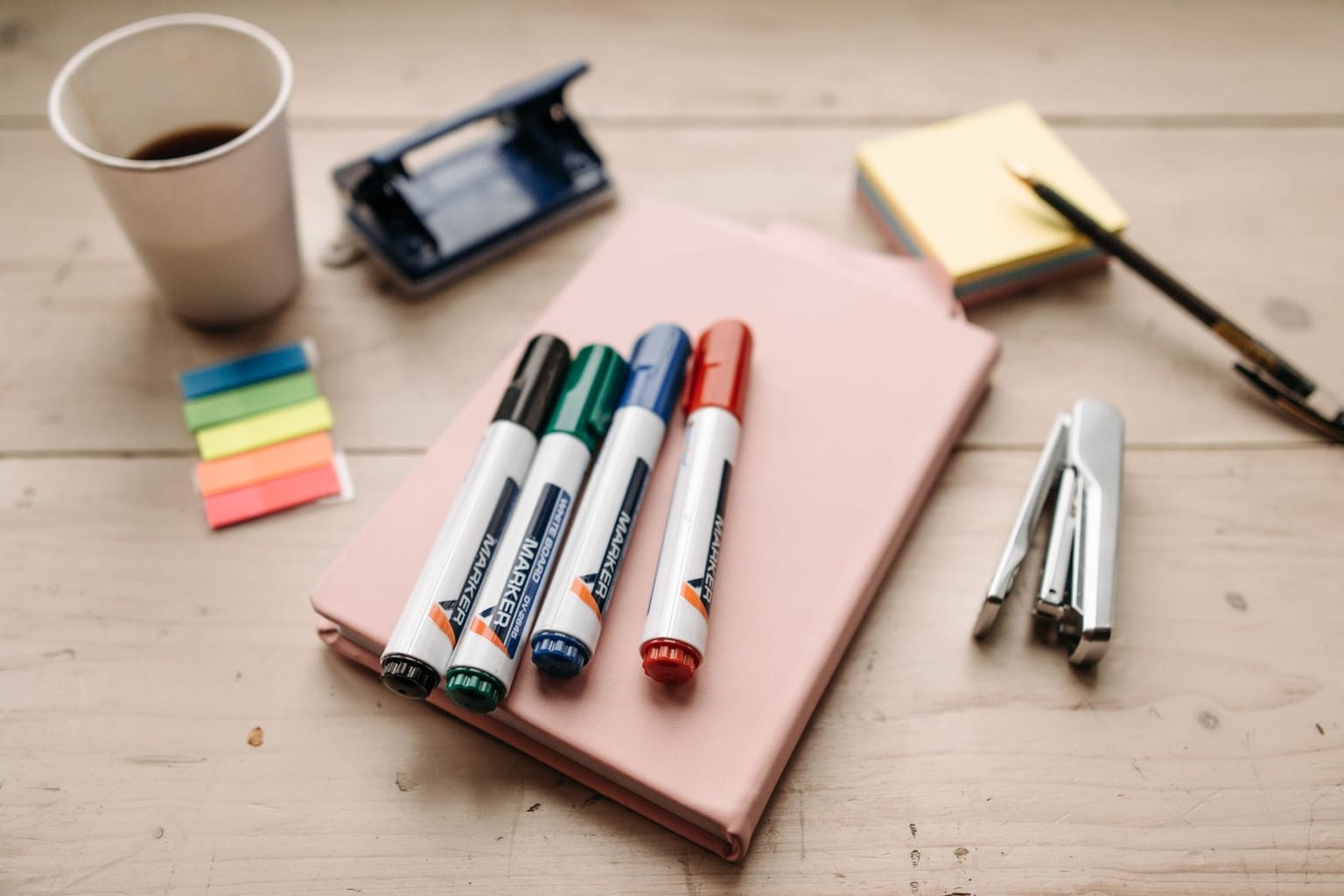 A teacher's desk with classroom supplies laid out: colored markers on a notebook, a pad of sticky notes, a pen, a stapler and a hole punch