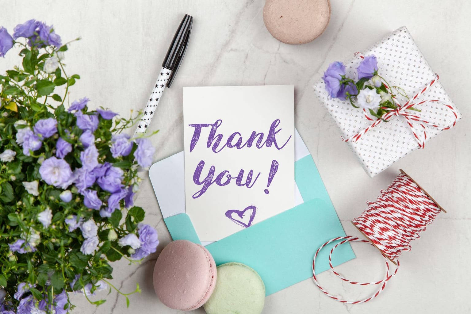 A thank-you card standing in an envelope on a desk beside a pen, a small potted plant and a wrapped gift, ready to be written