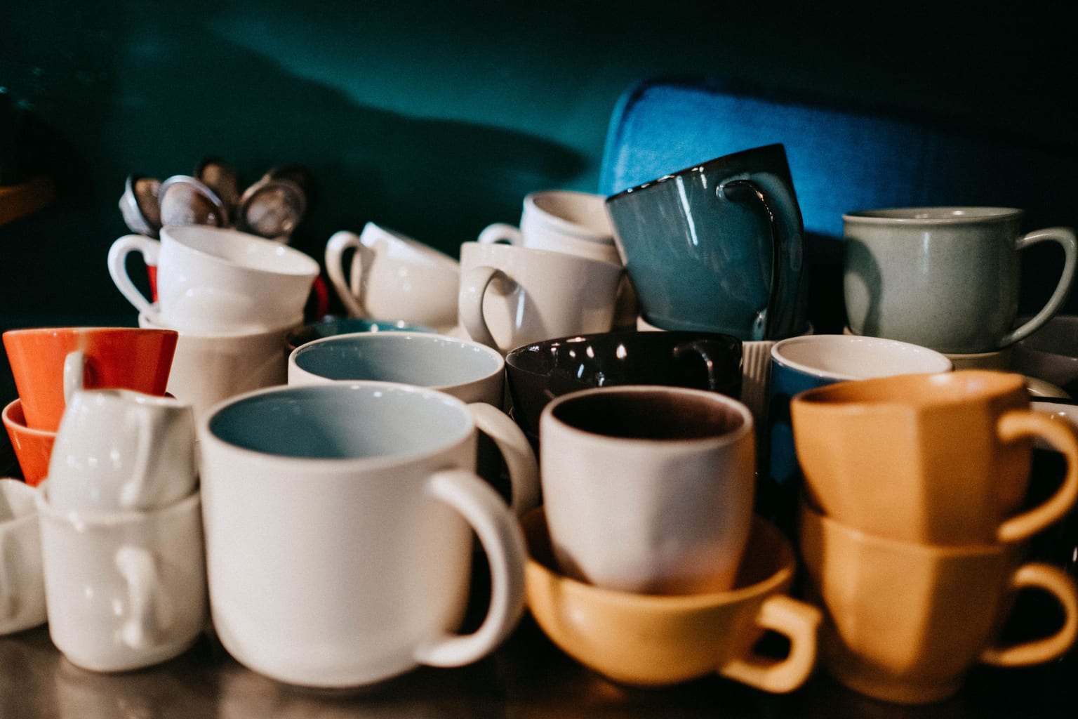 Around two dozen mismatched mugs stacked and crowded together on a kitchen counter, no two the same, the way a cupboard looks after several years of teacher gifts